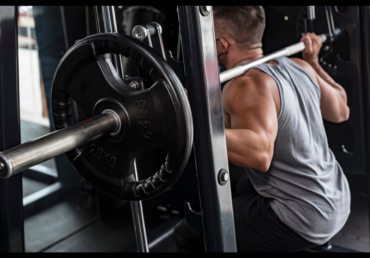 A man is doing a leg workout on a Smith machine in a gym. He is doing a squat with a barbell on his shoulders, loaded with weight. The essence of the photo is a close-up of a barbell with a manly back and arms. The weight plates are marked at 20 kg (44 lbs). The gist is made in a dark gym in order to show the exercise equipment.